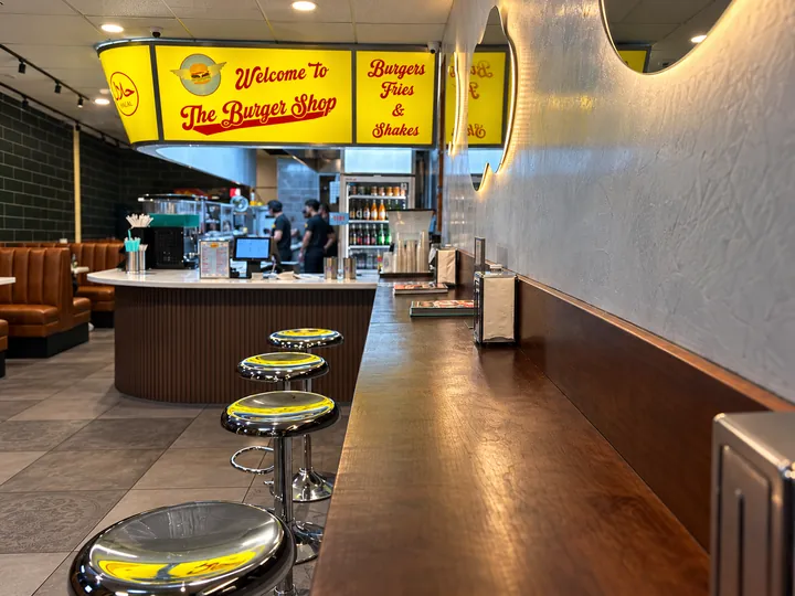 Chrome stools along the long wood counter, under the glowing yellow Welcome to The Burger Shop sign